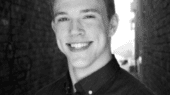 Black and white photo of a young man with short hair, smiling and wearing a button-up shirt, standing between two brick walls.