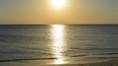 The sun sets over a calm ocean, casting a bright reflection on the water's surface and illuminating the sandy beach in the foreground.