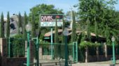 Outdoor view of a restaurant named "Rivas" with lush greenery, brick and green metal fence, and a sign indicating it is open every day. .