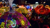 A colorful Mardi Gras umbrella decorated with painted masks and feathers is held up among a lively crowd during a nighttime parade.