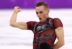 A figure skater in a maroon and black costume performs with an intense expression, raising a clenched fist during a routine on the ice.