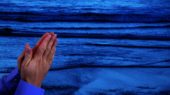 Two hands in a praying position in front of a blue-tinted ocean with gentle waves.