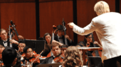 A conductor in a white jacket leads an orchestra performing on a stage, with musicians playing string instruments.