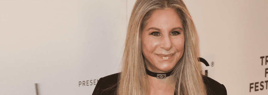 A person with long blonde hair, wearing a black outfit and choker, stands against a white backdrop at an event.