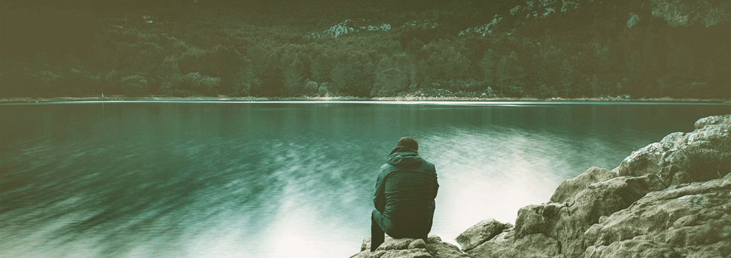 A person wearing a dark jacket sits on rocky terrain, facing a still body of water with a forested area in the background.