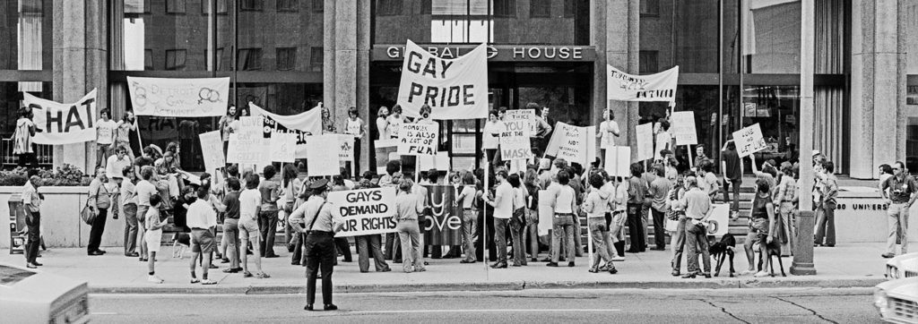 Black-and-white photo of a large group of protesters outside a building with signs and banners advocating for gay rights and demanding equality. One banner prominently reads "GAY PRIDE.
