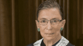 A person with glasses and green earrings is wearing a black outfit with a white lace collar. The background features dark wood paneling.