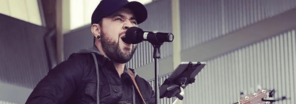 A man wearing a cap and a jacket is singing passionately into a microphone while playing an acoustic guitar on stage.