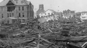 A scene of extensive destruction with debris and collapsed buildings in an urban area, including a partially standing large brick building and scattered remnants of wooden structures.