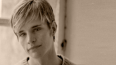 A sepia-toned portrait of a person with short hair, wearing a sweater, looking at the camera, with a window in the background.