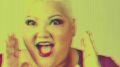 A person with short blond hair and red lipstick is holding their hands near their face, appearing surprised or excited, with a colorful lighting effect on the image.