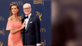 A man and woman stand together on the red carpet at an event, with a backdrop featuring "Creative Arts Emmys" and "Television Academy" logos. The man wears a suit and bow tie; the woman wears an off-shoulder dress.