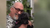 A man with glasses and a beard smiles while holding a small black dog in an outdoor setting with plants and lattice in the background.