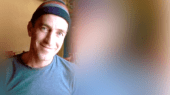 A man in a black beanie and gray T-shirt smiles while looking at the camera, with a blurred background.