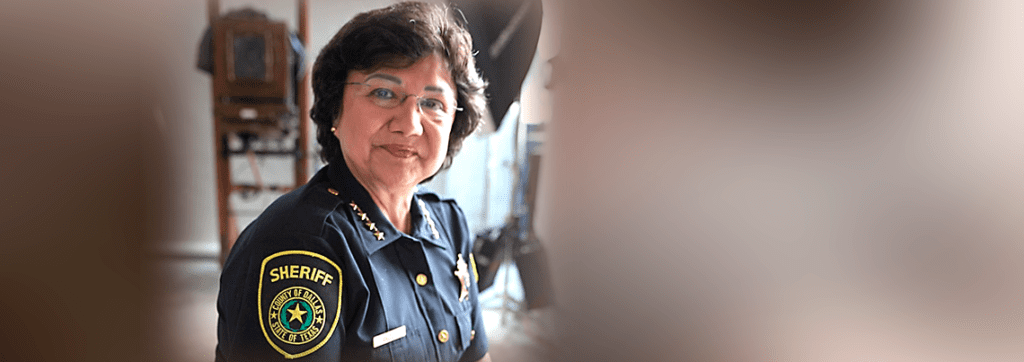 A sheriff in uniform with a badge and shoulder patch stands indoors, looking at the camera with a neutral expression.