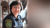 A sheriff in uniform with a badge and shoulder patch stands indoors, looking at the camera with a neutral expression.