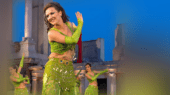 A woman in a green costume performs a dance outdoors, with ancient ruins in the background and two other dancers partially visible.