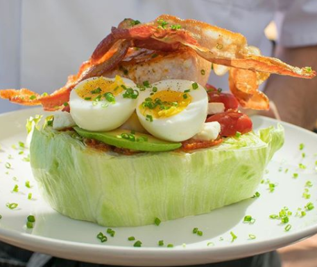 A wedge of iceberg lettuce topped with bacon strips, halved boiled eggs, tomato slices, avocado slices, and sprinkled with chopped chives sits on a white plate.