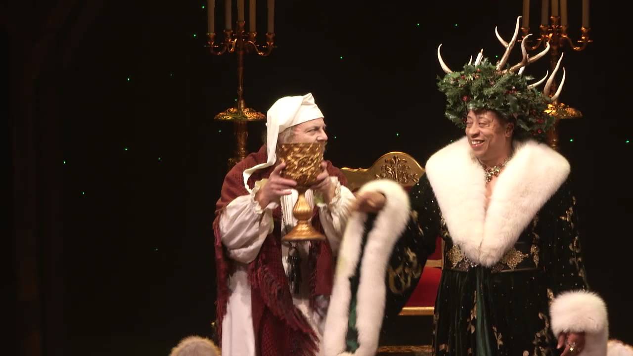 Two actors in elaborate costumes perform on stage. One wears a wreath with antlers and fur trim, while the other holds a large goblet. Both appear to be in a festive setting.