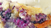 Close-up of a slice of blueberry cobbler topped with a scoop of vanilla ice cream. The cobbler has a golden, crumbly crust and visible juicy blueberries.