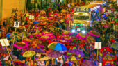 A large crowd of people with colorful umbrellas participate in a street parade at night, featuring a decorative bus with a "DANCE" sign.