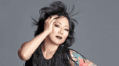 A person with dark hair and tattoos on their left arm holds their head with their left hand, wearing a fringed black outfit and dark makeup, against a gray background.