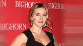 A woman in a black dress stands in front of a red backdrop with the word "BIGHORN" printed on it multiple times. She has blonde hair styled in an updo and is wearing drop earrings.