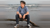 A person with short hair sits on a log at the beach, wearing a blue short-sleeve shirt, jeans, and sneakers. The ocean and waves are visible in the background.