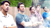 Seven men stand with arms crossed in front of a wooden fence, looking serious. Smoke wafts in front of them, suggesting a barbecue. Trees and bushes are visible in the background.