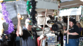 A group of people, many wearing masks and holding festive signs, participate in a celebration inside a building. The signs read "Mardi Gras" and "United Charities." The event includes decorations and beads.