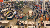 A crowded indoor trade show with numerous booths, showcasing various products including hot tubs, pools, and a large cow mascot in the center. Attendees are walking and interacting with exhibitors.