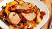 A plate of pork loin medallions topped with herbs, accompanied by roasted vegetables including squash and potatoes, with a fork and knife on a wooden table.