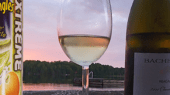 A can of Pringles next to a wine glass and a bottle of wine are placed on a surface, with a lakeside view and trees in the background at sunset.