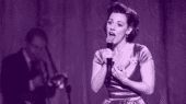 Woman in a vintage dress sings passionately into a microphone with a musician playing an instrument in the background. The image has a purple tint.