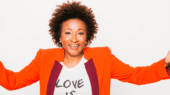 Person smiling with arms outstretched, wearing an orange blazer over a white t-shirt that reads "LOVE IS." Against a plain white background.