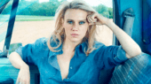 A person with blonde hair sits in the driver's seat of a vehicle, wearing a blue denim shirt. The background shows a field and trees. The person appears to be looking at the camera with a serious expression.