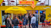 People gather under yellow umbrellas at "Sabor Latino" food stand at an outdoor venue. Bright yellow walls and signage, red chairs and tables are visible. Some people hold umbrellas while others chat.