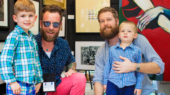 Two adults and two children smile at an art gallery. The two adults have beards and wear casual clothes, while the children wear plaid shirts. They are surrounded by framed artwork on the wall.