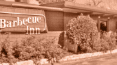 Exterior of a building with a sign that reads "Barbecue Inn". The structure has a low roof and is surrounded by neatly trimmed bushes.