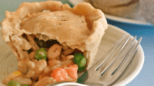 A small vegetable pot pie with a flaky crust sits partially cut open on a white plate next to a fork. Peas, carrots, corn, and beans are visible inside the pie.