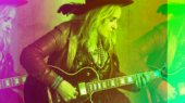 A person with long hair wearing a hat and a leather jacket plays an electric guitar. The image has a colorful, layered effect with pink, green, and yellow hues.