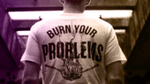 A person wearing a white t-shirt with the text "Burn Your Problems" featuring an illustration of a flame. The person is standing in a dimly lit urban environment.