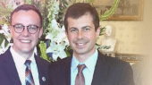 Two men in suits smile for a photo in front of a floral arrangement and a bust sculpture.