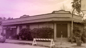 A brick building with a tile roof, surrounded by trees and bushes, is closed off with a barrier. A parked car is visible on the left.