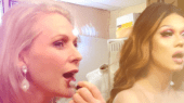 Two people apply lipstick in front of mirrors. The person on the left has blonde hair and is using a lipstick tube, while the person on the right has long brown hair and applies lip gloss.