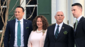 Four individuals stand in front of a white building with green shutters, all dressed in formal attire, including suits and a dress.