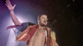 Shirtless man draped in a rainbow flag sings into a microphone on a dimly lit stage.