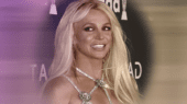 A person with long blonde hair, wearing a dress with a glittering star-shaped decoration, smiles while standing in front of a backdrop with text and logos.