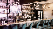 A modern, well-lit bar interior with numerous bottles displayed on shelves, plush blue bar stools, hanging light fixtures, and a TV screen behind the counter.