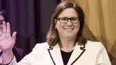 A woman wearing glasses and a white blazer with black trim waves, standing next to others in a formal setting.